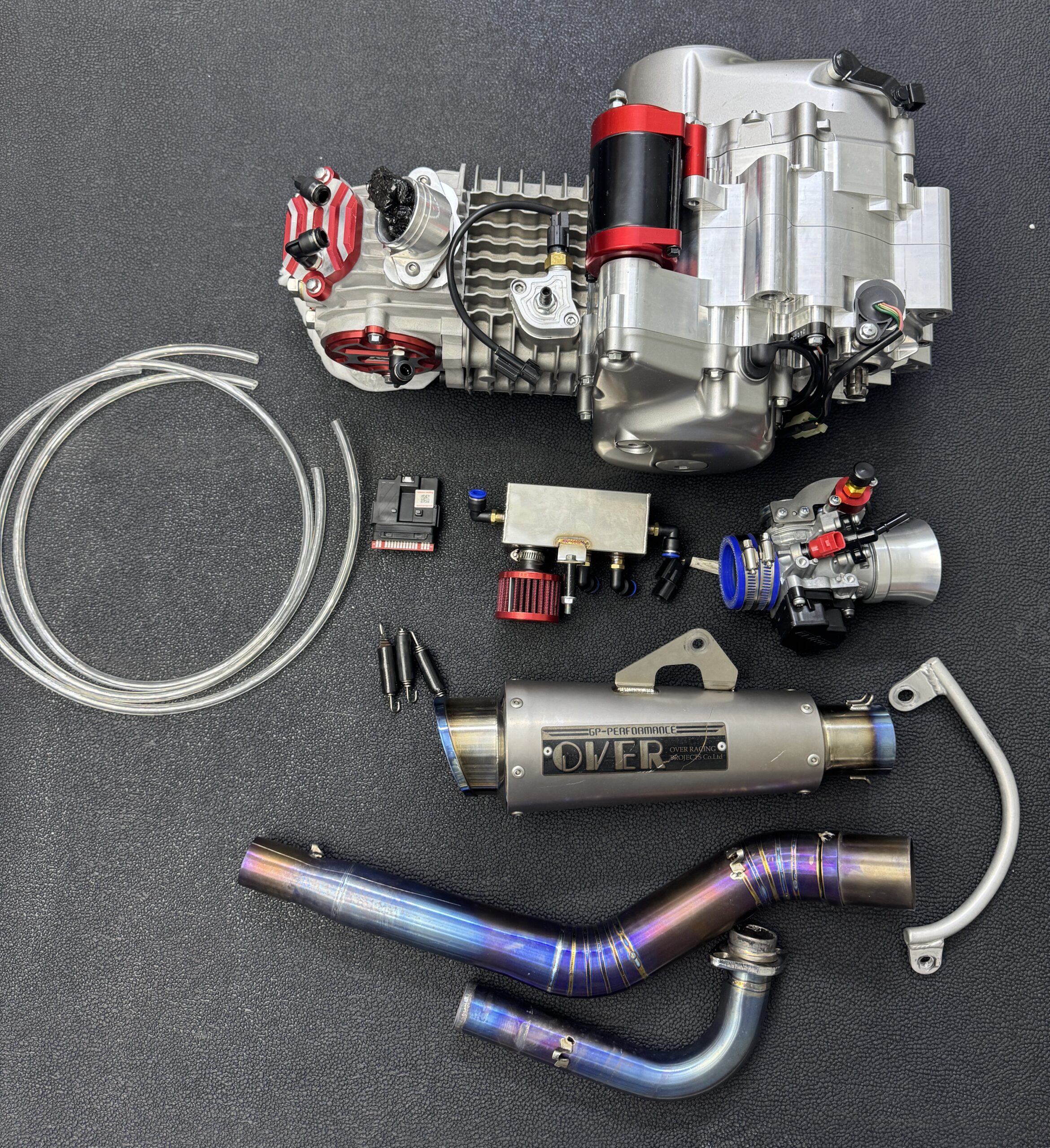 High-performance engine tuning and upgrade kit featuring a Monkey mini motorcycle engine, performance exhaust pipe, ECU, throttle body, wiring, and tuning accessories laid out on a black surface.