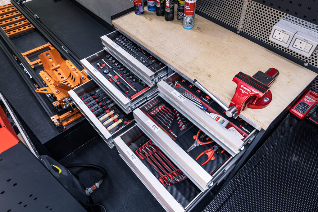 An organized motorcycle repair workstation with open tool drawers containing neatly arranged hand tools, including screwdrivers, ratchets, spanners, and pliers, next to a mounted bench vise and motorcycle assembly equipment.