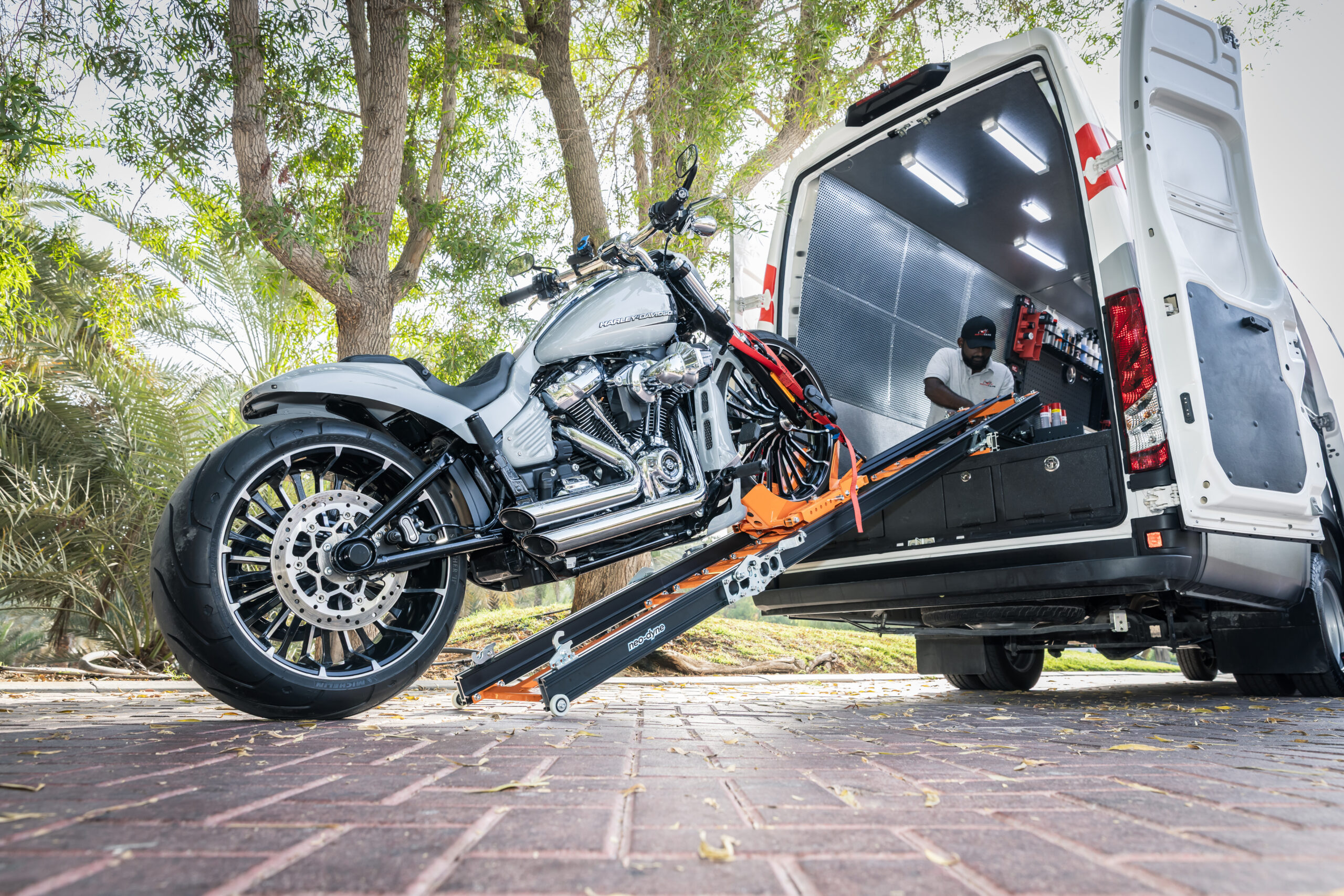 A silver Harley-Davidson motorcycle is being carefully loaded into the back of a specialized Motocare service van using a sturdy loading ramp. The van is equipped with bright interior lights, organized storage cabinets, and professional-grade tools, all set in a tree-lined outdoor environment.